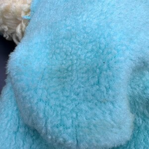 Peut inclure: Gros plan sur un tissu bleu clair, duveteux et textur&eacute;. La mati&egrave;re semble douce et moelleuse, avec un poil dense et boucl&eacute;. Une petite section de tissu blanc cass&eacute; et boucl&eacute; est visible dans le coin sup&eacute;rieur gauche.