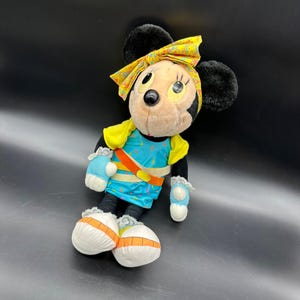 Peut inclure: Un jouet en peluche de Minnie Mouse portant une tenue jaune et bleue avec un bandeau jaune. Le jouet est assis sur une surface noire.