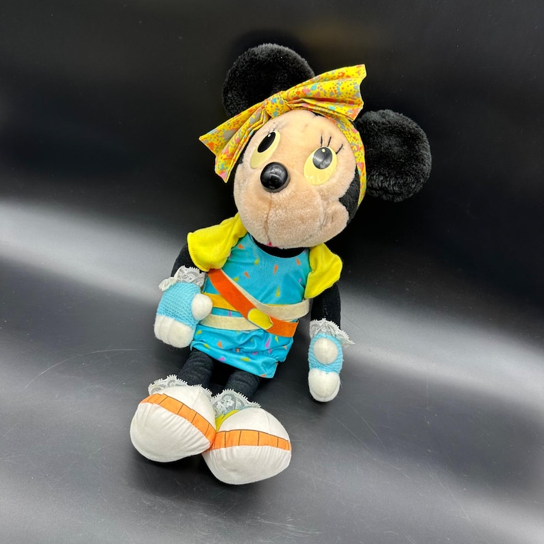 Peut inclure: Un jouet en peluche de Minnie Mouse portant une tenue jaune et bleue avec un bandeau jaune. Le jouet est assis sur une surface noire.