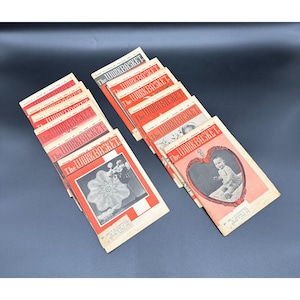 Może przedstawiać: Kolekcja starych magazynów "The Workbasket". Okładki są czerwono-białe z tytułem magazynu pogrubioną czcionką. Niektóre okładki przedstawiają zdjęcia dziecka, serwetki i inne ilustracje. Magazyny są ułożone na ciemnej powierzchni.