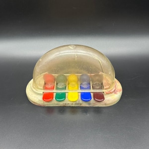 May include: Vintage Tap-A-Toy musical toy with a clear plastic dome and a base with colorful keys. The toy features five colored keys and five matching colored pegs. The text "TAP-A-TOY" is printed on the base.