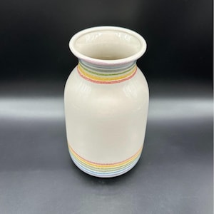May include: A cream-colored ceramic vase with a rounded body and a flared rim. The vase features horizontal bands of thin, textured stripes in pastel shades of pink, yellow, green, and blue encircling the neck and the base of the vessel.