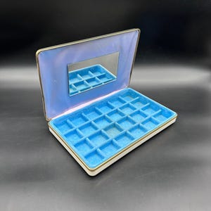 May include: Open vintage jewelry box with a blue velvet interior and a small mirror. The box has a white exterior with gold trim and a blue satin lining. The interior is divided into small compartments for storing jewelry.