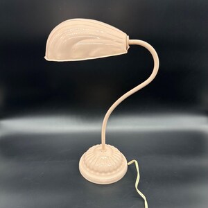 K&ouml;nnte beinhalten: Eine Vintage-Schreibtischlampe in einem zarten Rosa. Die Lampe hat ein gebogenes Schwanenhals-Design mit einem abgerundeten Schirm und einem dekorativen Sockel. Der Schirm hat eine gerippte Textur, und der Sockel ist kreisf&ouml;rmig mit einem floralen Muster. Ein cremefarbenes Kabel erstreckt sich vom Sockel.