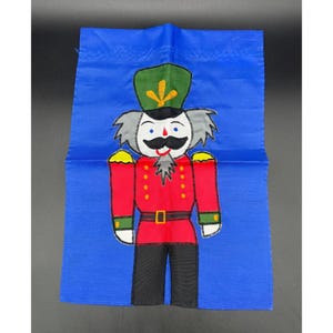 Puede incluir: Una bandera de jardín de tela azul con un diseño de cascanueces de dibujos animados. El cascanueces lleva un uniforme rojo y dorado con un sombrero verde y una barba blanca.