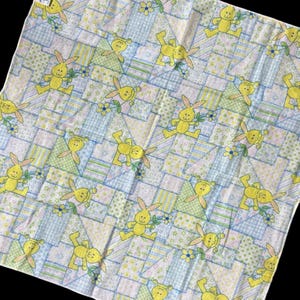 Peut inclure: Un tissu carré de style patchwork avec un motif répétitif de lapins de dessin animé jaunes et de petites fleurs bleues. Le fond est un mélange de carrés bleu clair, verts et blancs.
