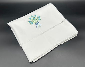May include: White linen pillowcase with an embroidered bouquet of blue flowers and green leaves tied with a blue ribbon. The pillowcase has a decorative stitched border and is folded neatly.