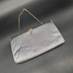 May include: A rectangular silver clutch bag with a textured metallic finish. The bag features a thin silver-tone metal chain strap and a central decorative clasp with a leaf-like shape. The frame is constructed from silver-tone metal hardware.
