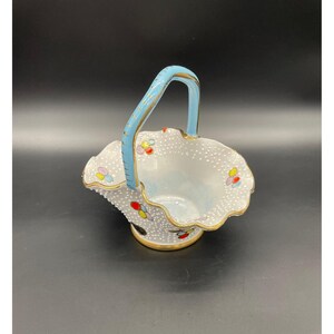 May include: A small white ceramic basket with a blue handle and colourful floral accents. The basket has a scalloped edge and a dotted pattern.