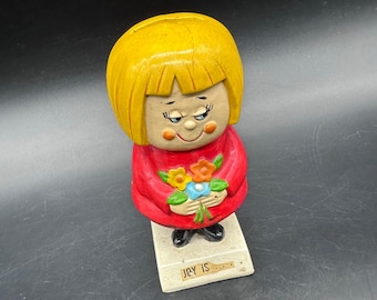 May include: A vintage ceramic figurine of a girl with yellow hair and a red dress, holding a bouquet of flowers. The figurine has a smiling face and the text "JOY IS" on the base. The figurine is a collectible item.