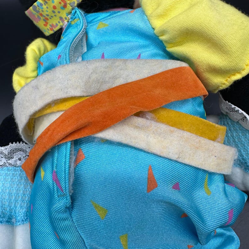Peut inclure: Un jouet en peluche bleu et jaune avec une fermeture &eacute;clair et une ceinture en tissu blanche, orange et jaune. Le jouet a un motif de confettis.