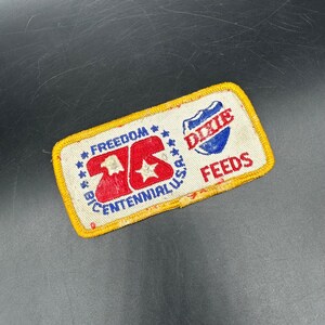 Puede incluir: Parche rectangular con borde amarillo. El parche presenta el texto "FREEDOM" y "BIG CENTENNIAL U.S.A." en azul, y "DIXIE FEEDS" en rojo y azul. El centro tiene un "26" rojo con una estrella y un águila blancos.