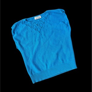 May include: A bright turquoise knit top with a ribbed hem and a decorative diamond pattern around the neckline. The top has short sleeves and a scoop neck. The label reads "Faith Kniits".