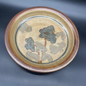 May include: A round ceramic plate with a floral design. The plate features a brown rim, a tan center, and a blue and gray floral pattern. The plate is suitable for serving food or as a decorative piece.
