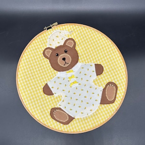 May include: A yellow and white gingham fabric hoop with a brown bear wearing a white nightgown with yellow flowers embroidered on it.