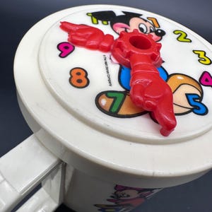 Puede incluir: Un reloj vintage de Mickey Mouse blanco con una manecilla roja en forma de mano. La esfera del reloj presenta a Mickey Mouse y n&uacute;meros coloridos. El asa es blanca. El reloj es un art&iacute;culo de colecci&oacute;n.
