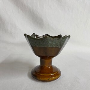 May include: A brown and green ceramic pedestal bowl with a scalloped rim. The bowl has a glazed finish and a pedestal base.