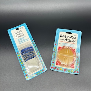 Puede incluir: Dos paquetes de suministros de costura. El paquete de la izquierda es azul y blanco y contiene un dedal de cuero. El paquete de la derecha es azul y amarillo y contiene cera de abejas y un soporte para el tejido de velas y el acolchado. El texto del paquete de la izquierda dice "Quilter's Leather Thimble" y el texto del paquete de la derecha dice "Beeswax and Holder".