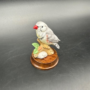 May include: A ceramic figurine of a bird with gray and white feathers, a red beak, and black eyes. The bird is perched on a brown branch with green leaves and sits on a round, wooden base. The figurine is a decorative item.