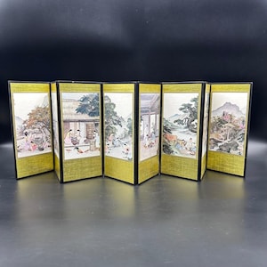 May include: A six-panel folding screen with gold borders and black frames. Each panel features a detailed, colorful illustration of a traditional Asian landscape with figures, trees, and buildings. The screen is displayed on a dark surface.