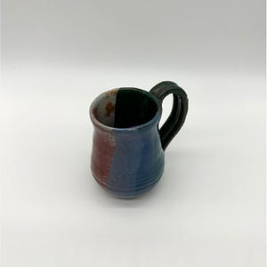 May include: A small, handmade ceramic mug with a black handle. The mug is decorated with a blue, green, and red glaze.