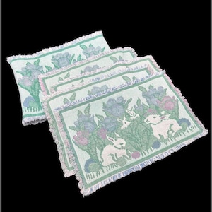 May include: A set of four rectangular placemats with a light green background. Each placemat features a floral and bunny design in shades of blue, pink, and white. The edges are fringed.