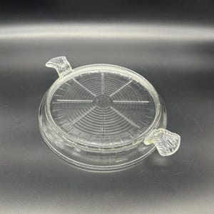 May include: A clear glass hot pad with two handles. The hot pad has a circular design with concentric circles.