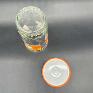 May include: Clear glass jar with a textured, geometric design. The jar has an orange and white label and a matching lid. The lid is white with an orange rim. The jar is empty and sitting on a gray surface.