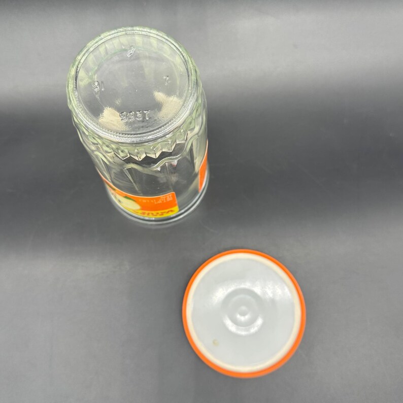 May include: Clear glass jar with a textured, geometric design. The jar has an orange and white label and a matching lid. The lid is white with an orange rim. The jar is empty and sitting on a gray surface.
