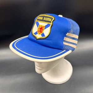 May include: A blue trucker hat featuring a white mesh back and a front patch with the text NOVA SCOTIA above a blue saltire and a red lion emblem. The hat has a flat brim with a white trim and three horizontal white stripes on the side mesh.