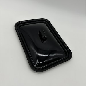 以下が含まれることがあります： 小さな湾曲したハンドルが付いた、長方形の光沢のある黒い蓋。蓋には丸みを帯びた隆起したエッジと滑らかで反射性の表面があります。このオブジェクトは、容器または調理器具の蓋である可能性があります。