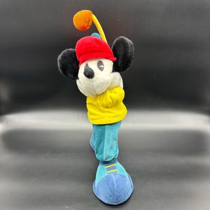 Puede incluir: Un mu&ntilde;eco de peluche de Mickey Mouse con un sombrero rojo, una camisa amarilla y pantalones azules. Est&aacute; sosteniendo un palo de golf y est&aacute; en una postura de golf.