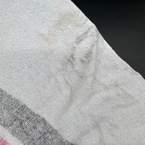 Op de afbeelding: Close-up van een witte handdoek met grijze en roze strepen. De handdoek vertoont vlekken en slijtage. De stof is een zacht, absorberend materiaal, typisch voor badhanddoeken.