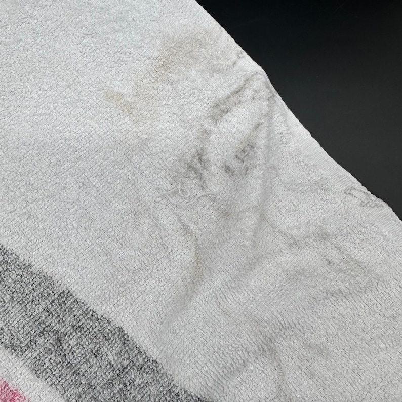 Op de afbeelding: Close-up van een witte handdoek met grijze en roze strepen. De handdoek vertoont vlekken en slijtage. De stof is een zacht, absorberend materiaal, typisch voor badhanddoeken.