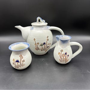 May include: A white ceramic teapot, creamer, and sugar bowl set with a mushroom design. The teapot has a blue and white glaze, while the creamer and sugar bowl have a blue and white glaze with a brown rim.