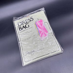 Puede incluir: Una funda de vestido de plástico transparente con el texto "DRESS BAG a protective cover". La funda mide 122 cm de largo, 61 cm de ancho y 8 cm de profundidad. Presenta un gráfico de un vestido en el interior. La funda protege contra el polvo, las polillas y el moho.