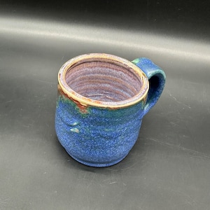 May include: A blue ceramic mug with a speckled glaze and a light brown rim. The inside of the mug has a purple and white swirl pattern.