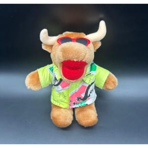 May include: A plush toy bull with a brown body, white horns, and red sunglasses. It wears a light green shirt with a colorful design. The bull has a large red tongue sticking out.