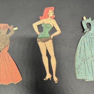 Puede incluir: Tres recortes de mu&ntilde;ecas de papel vintage sobre un fondo negro. Los recortes representan a una mujer con cors&eacute; y pantalones cortos verdes, y dos vestidos en tonos coral y verde azulado. La mujer tiene el pelo rojo y est&aacute; en una pose con las manos en las caderas.
