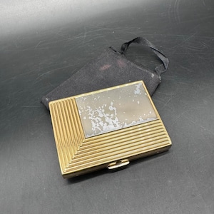 May include: A vintage gold-toned compact with a rectangular mirror and a black fabric pouch. The compact features a geometric design with vertical lines and a clasp closure. The mirror shows signs of wear.