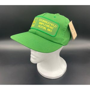 May include: A green baseball cap with a curved brim. The cap features a rectangular patch with the text "STUBBLEFIELD IMPLEMENT ALTON, MO" in yellow lettering. A small tag is attached to the side.