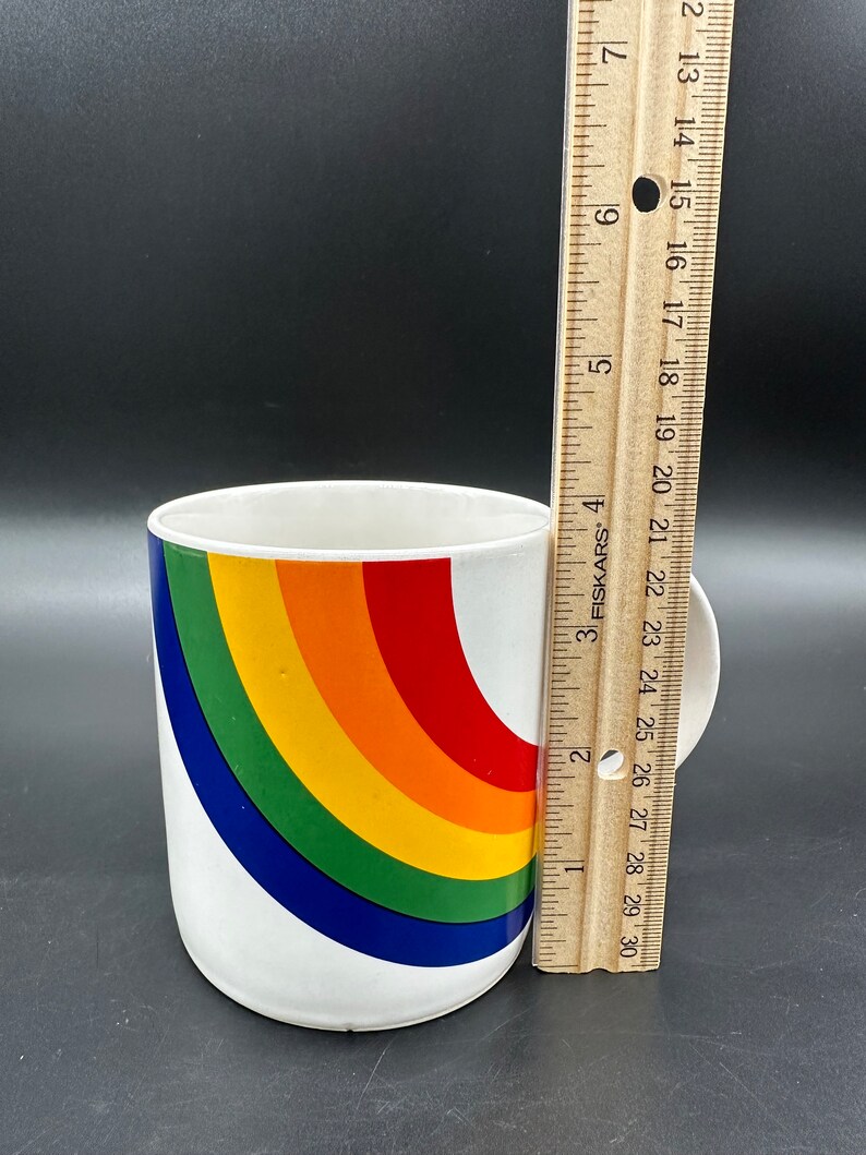 May include: White ceramic mug with a rainbow design. The rainbow is made up of red, orange, yellow, green, blue, and purple stripes. The mug is about 4 inches tall.