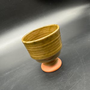May include: A handmade ceramic goblet with a textured, earthy brown exterior and a reddish-orange base. The goblet has a wide, angled opening and a footed base, showcasing a rustic aesthetic. The piece is likely suitable for beverages or decorative use.