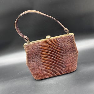 Puede incluir: Un bolso vintage marrón con un patrón texturizado similar al de un reptil. El bolso tiene un asa curva y un cierre dorado. El color del bolso es una mezcla de marrón y marrón rojizo.