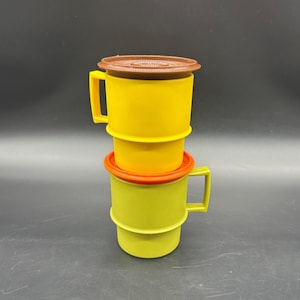 Puede incluir: Tres tazas de plástico de colores apiladas una encima de la otra. La taza superior es amarilla con una tapa marrón. La taza del medio es amarilla con una tapa naranja. La taza inferior es verde.