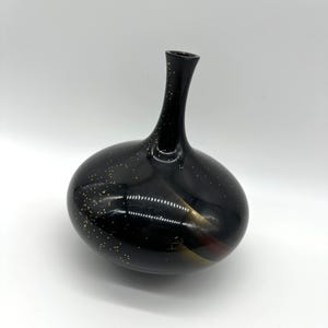 May include: A black glass vase with a rounded body and a slender neck, featuring a glossy finish and scattered gold-colored flecks across the surface. The design includes subtle amber-toned highlights that catch the light on the curved base.