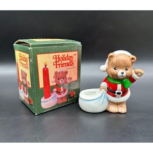 May include: A ceramic Holiday Friends candle holder featuring a brown bear dressed in a Santa Claus outfit, holding a blue sack. The candle holder is next to its original green box with the words "Holiday Friends" printed on it.
