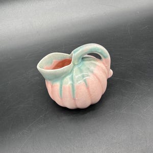May include: A small, ceramic creamer in the shape of a pumpkin. The creamer is pink with green and blue accents. It has a handle and a spout for pouring. The interior is a light pink color.