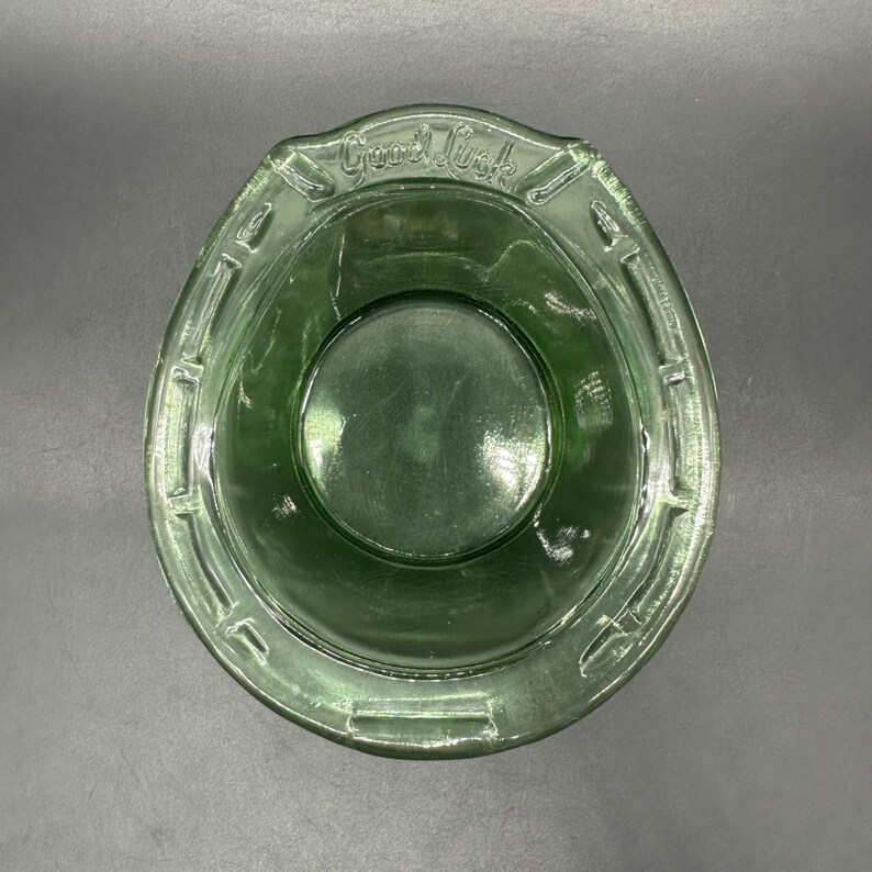 May include: A green glass bowl with a horseshoe shape and the words "Good Luck" embossed on the rim. The bowl has a circular base and a textured rim, perfect for serving snacks or as a decorative piece.