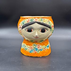 May include: Orange ceramic planter with a stylized face of a woman with black hair, large eyes, and a floral design around the head and neck.